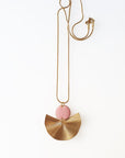 A necklace sits against a white background. It features a gold chain, a pink enamel connector dot and a textured brass fan piece.