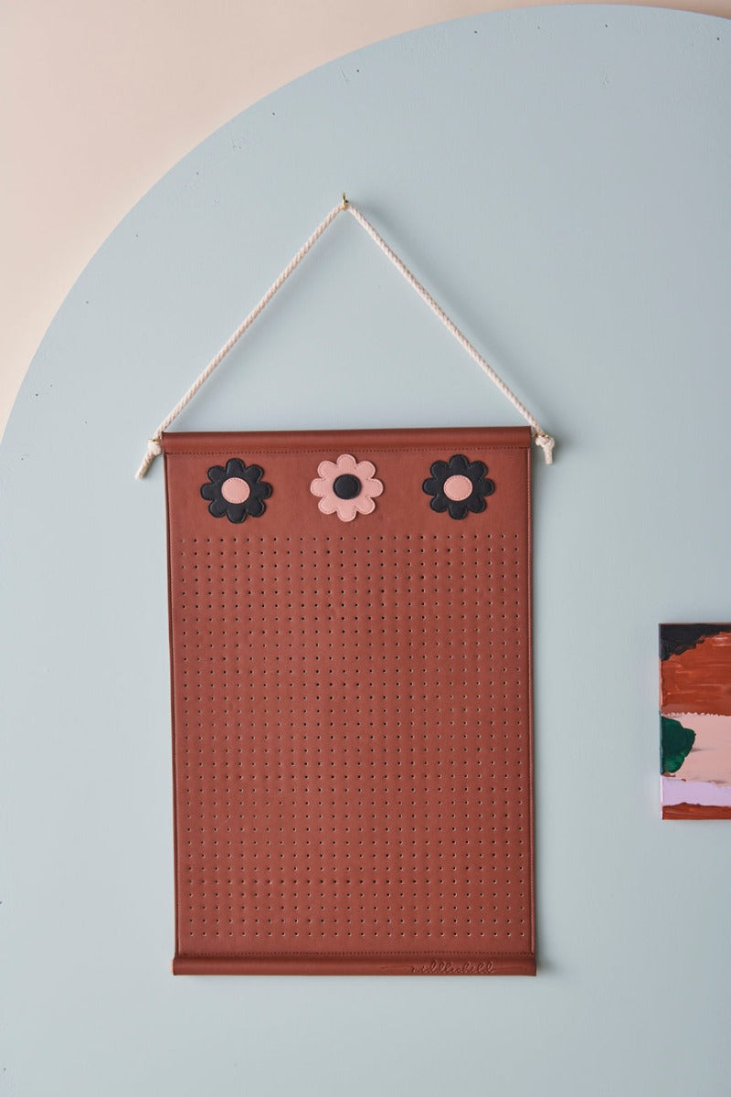 A brown wall hanger with three brown and pink flowers at the top is hung by a rope and styled against a blue arch and pink wall. Next to the wall hanger is a small abstract painting. The image shows a close up of the wall hanger with perforated holes for hanging jewellery and the word middlechild written in script across the bottom edge.