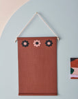 A brown wall hanger with three brown and pink flowers at the top is hung by a rope and styled against a blue arch and pink wall. Next to the wall hanger is a small abstract painting. The image shows a close up of the wall hanger with perforated holes for hanging jewellery and the word middlechild written in script across the bottom edge.
