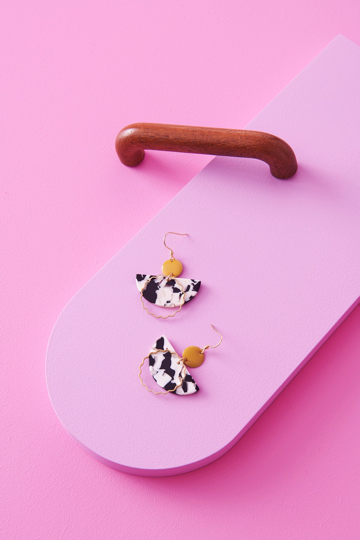 A pair of dangle earrings with a hook sit styled against a pink background next to a wooden drawer handle. The earrings feature a chartreuse enamel dot, a black and white D-shaped acrylic, and a wavy brass ring.