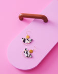 A pair of dangle earrings with a hook sit styled against a pink background next to a wooden drawer handle. The earrings feature a chartreuse enamel dot, a black and white D-shaped acrylic, and a wavy brass ring.