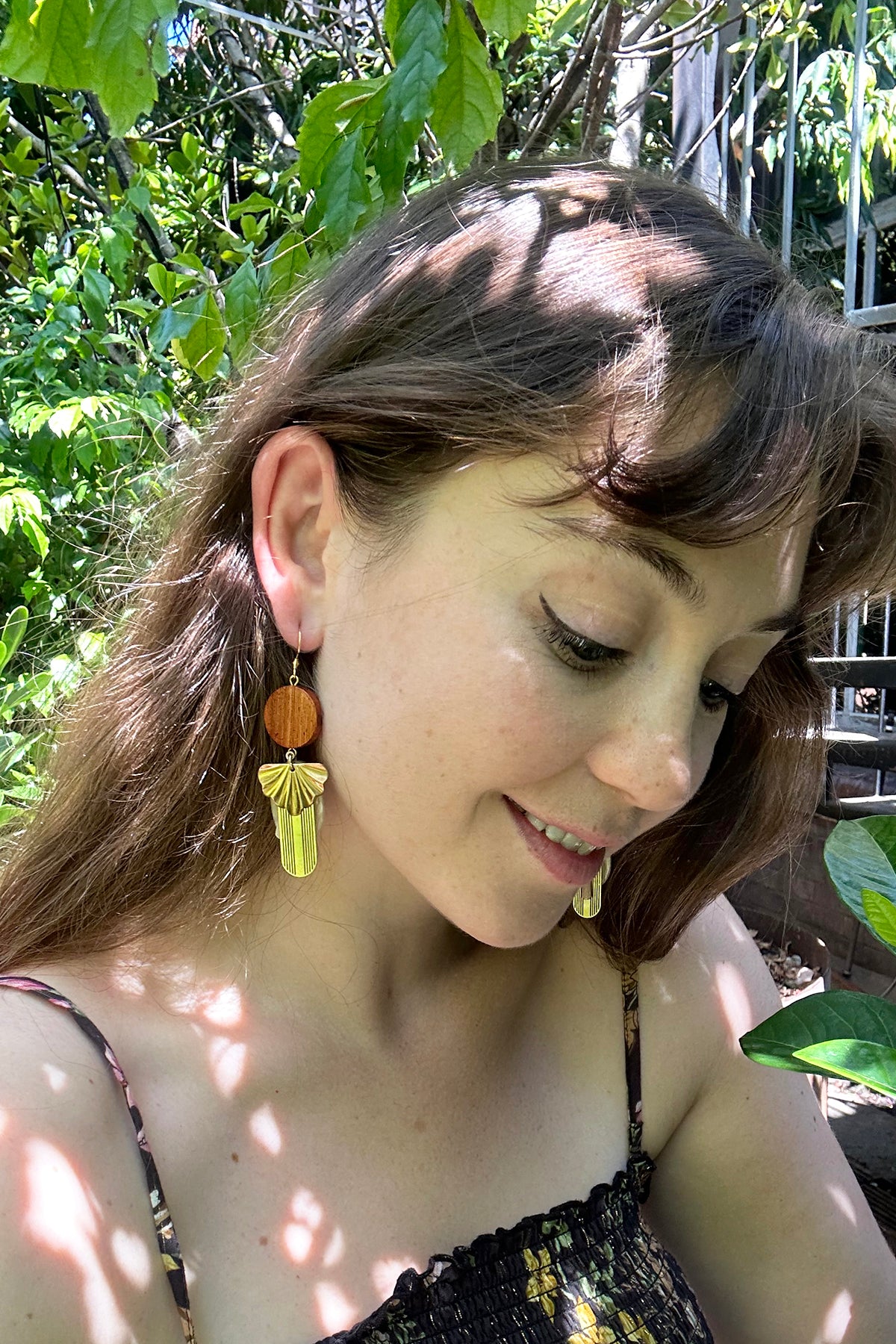 A lady with brown hair sits outside against a tree. She shows a sideview of the Heartsong earrings in Macadamia and wears a strappy floral top with rouching detail. 