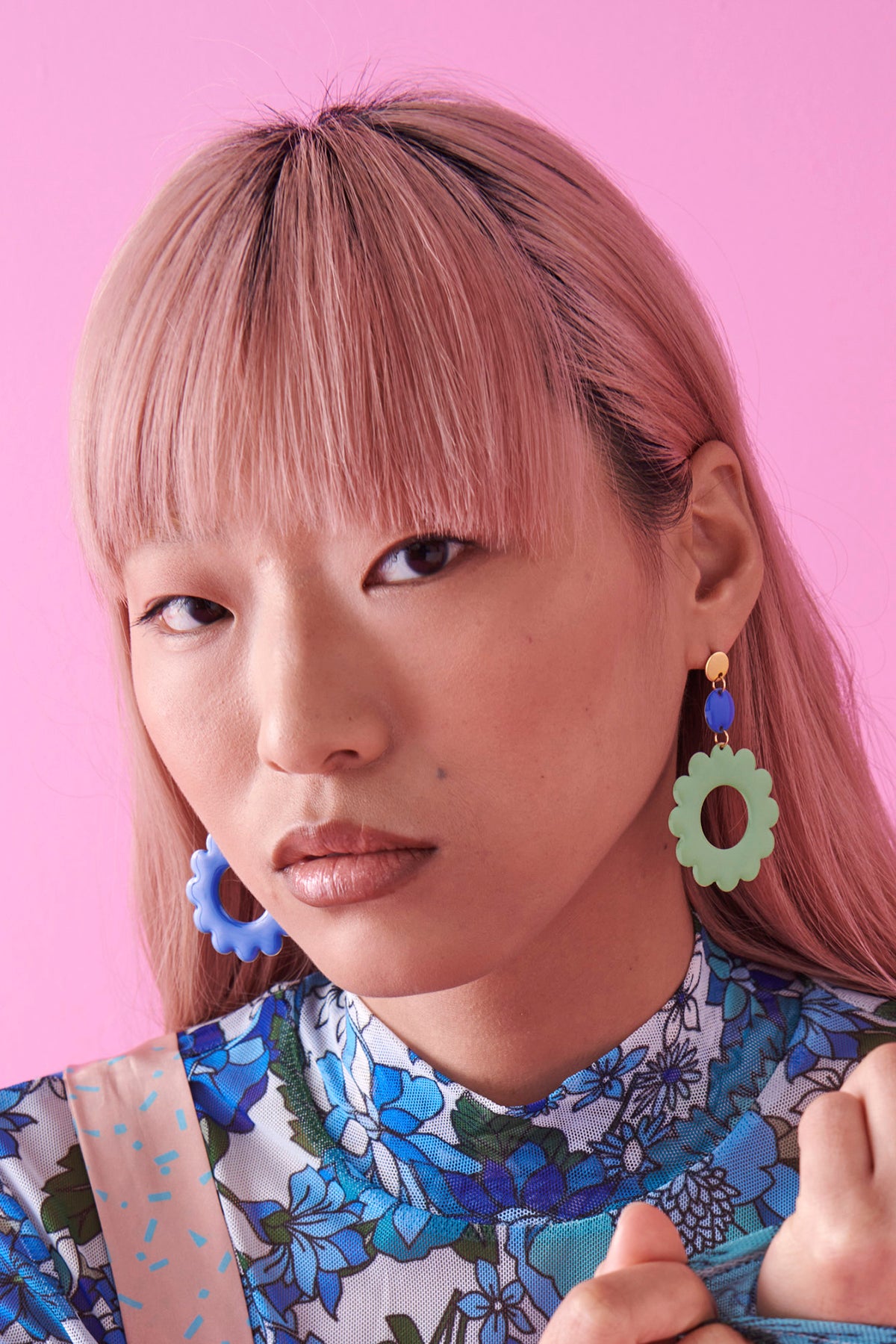 A lady with pink hair and a fringe sits against a bright pink background. She wears a blue floral top and has the Elton earrings in sapphire and sage on. Her head is turned slightly to make the sage flower on the Elton earring most visible. 
