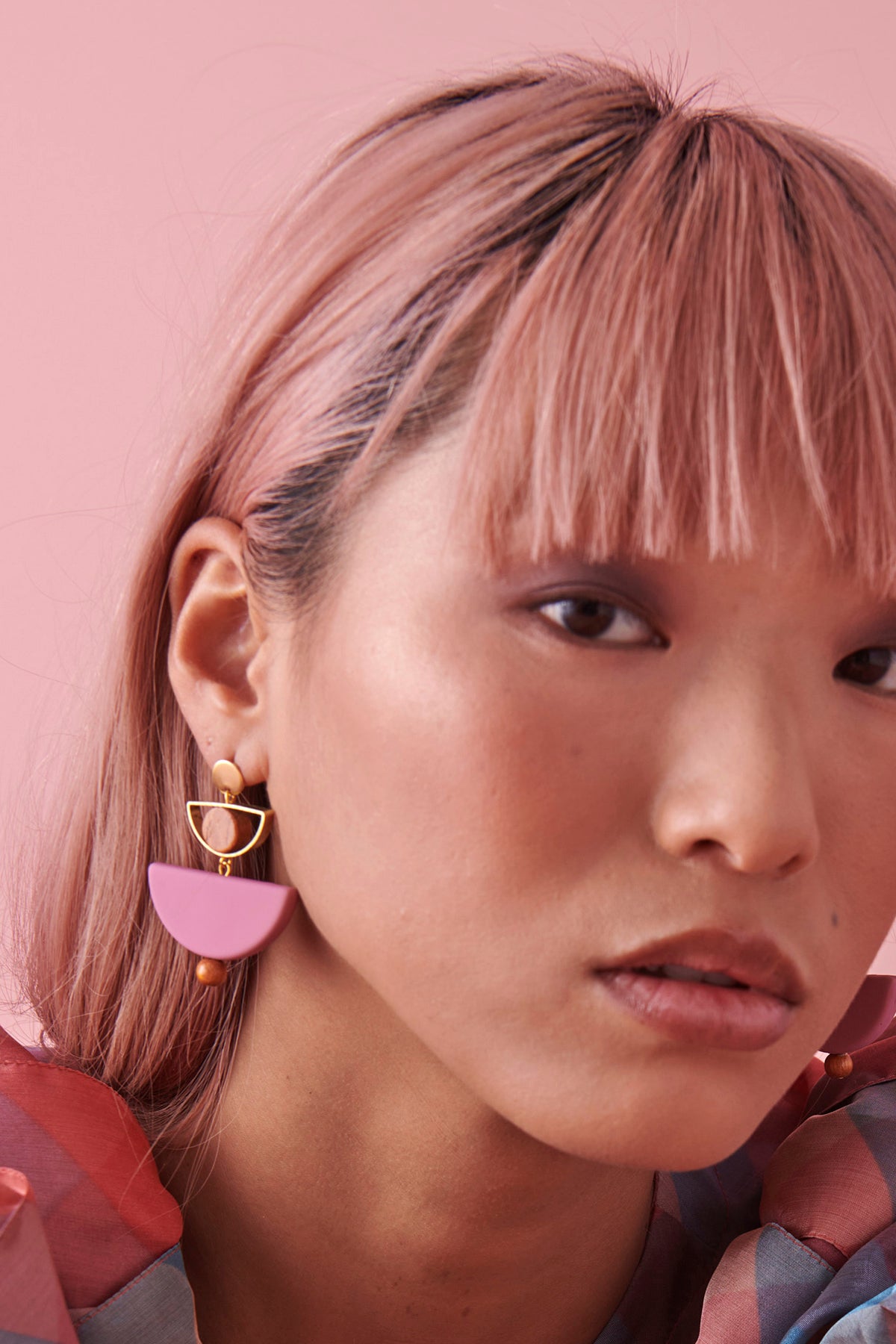 A closeup of a lady with pink hair against a pink background. She wears the  Mini Marcel earrings in grape and wears a pink and blue check  top