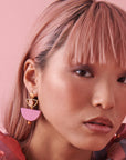 A closeup of a lady with pink hair against a pink background. She wears the  Mini Marcel earrings in grape and wears a pink and blue check  top