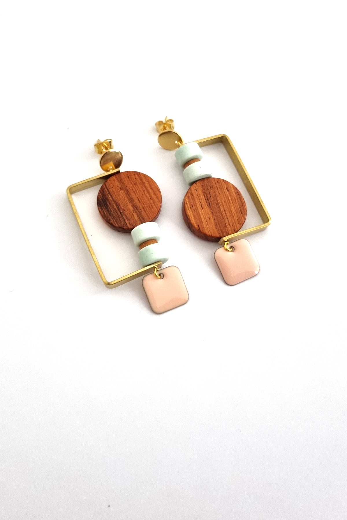 A pair of stud dangle earrings lay against a white background. They feature a half square brass shape that envelopes a circular wooden bead, and two aqua stone beads separated by a small wooden bead. The design is flipped in each ear and a peach square enamel dangle hangs from the bottom on each earring.