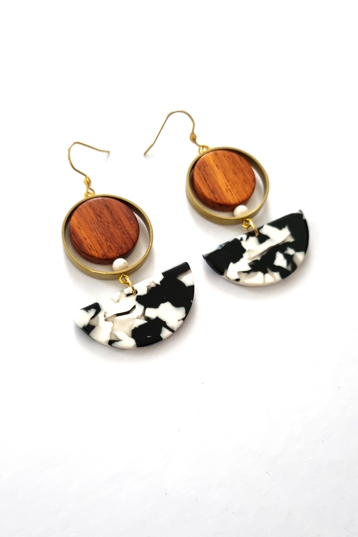 A pair of hook dangle earrings sit against a white background. They feature a circular wooden bead and a small white bead encircled by a brass ring. Below dangles a black and white D-shaped acrylic piece. 