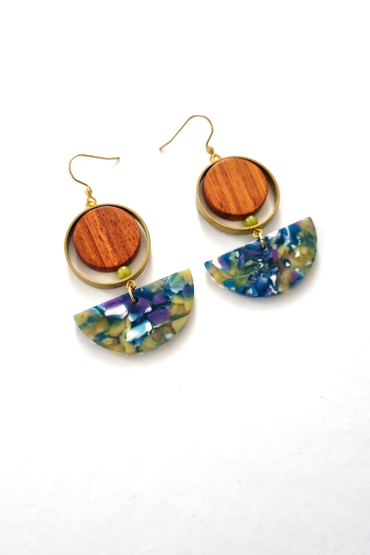 A pair of hook dangle earrings sit against a white background. They feature a circular wooden bead and a small green bead encircled by a brass ring. Below dangles a green and blue multicoloured D-shaped acrylic piece.