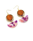 A pair of hook dangle earrings sit against a white background. They feature a circular wooden bead and a small purple bead encircled by a brass ring. Below dangles a pink and purple multicoloured D-shaped acrylic piece.