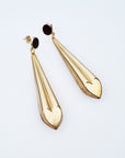 A pair of statement dangle earrings with a stud top sit against a white background. They feature a black enamel stud and a long brass drop with a heart shape at the bottom.