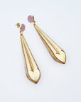 A pair of statement dangle earrings with a stud top sit against a white background. They feature a mauve enamel stud and a long brass drop with a heart shape at the bottom.