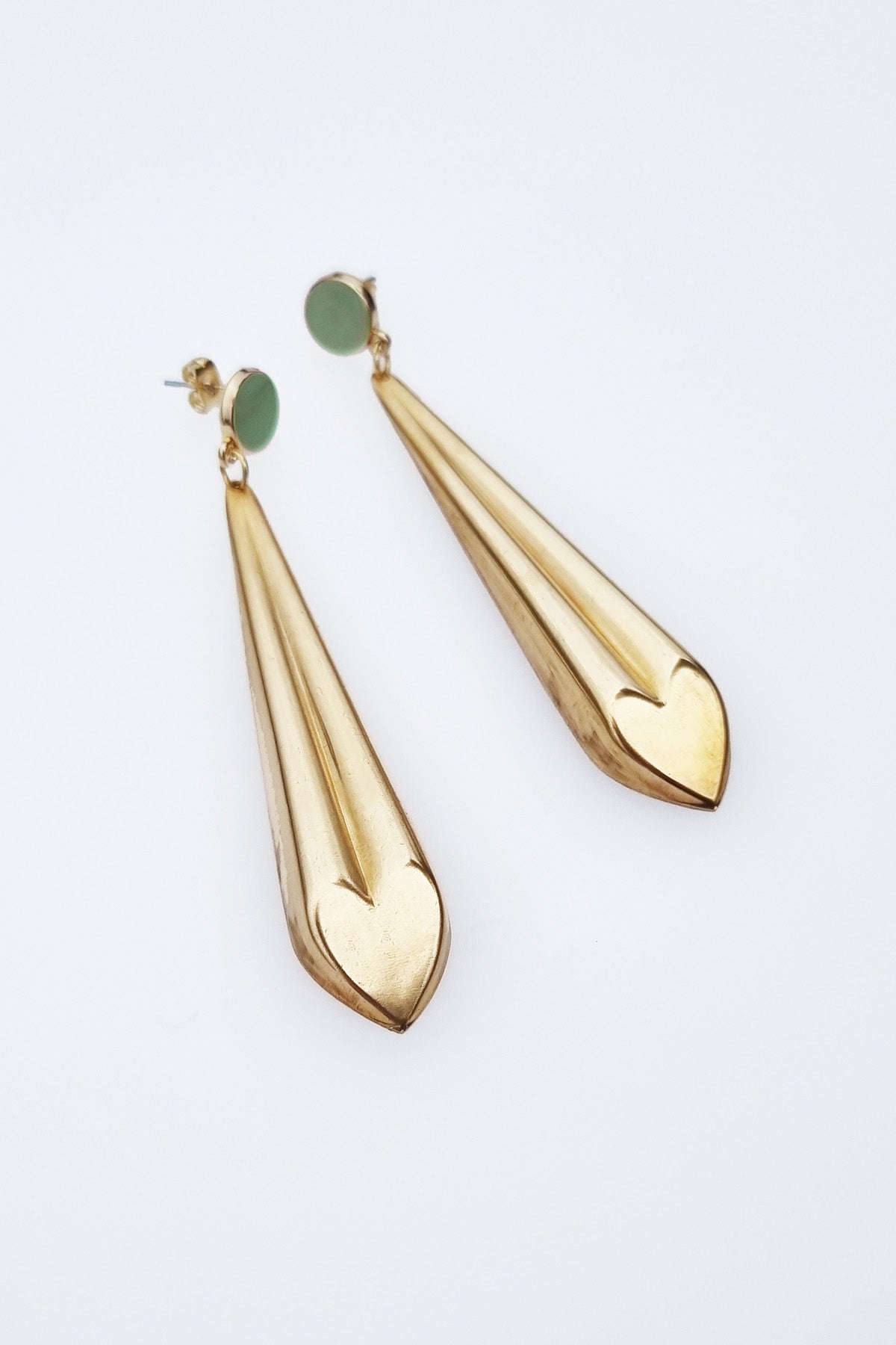A pair of statement dangle earrings with a stud top sit against a white background. They feature a sage enamel stud and a long brass drop with a heart shape at the bottom.