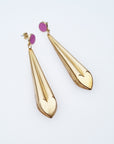 A pair of statement dangle earrings with a stud top sit against a white background. They feature a violet enamel stud and a long brass drop with a heart shape at the bottom.