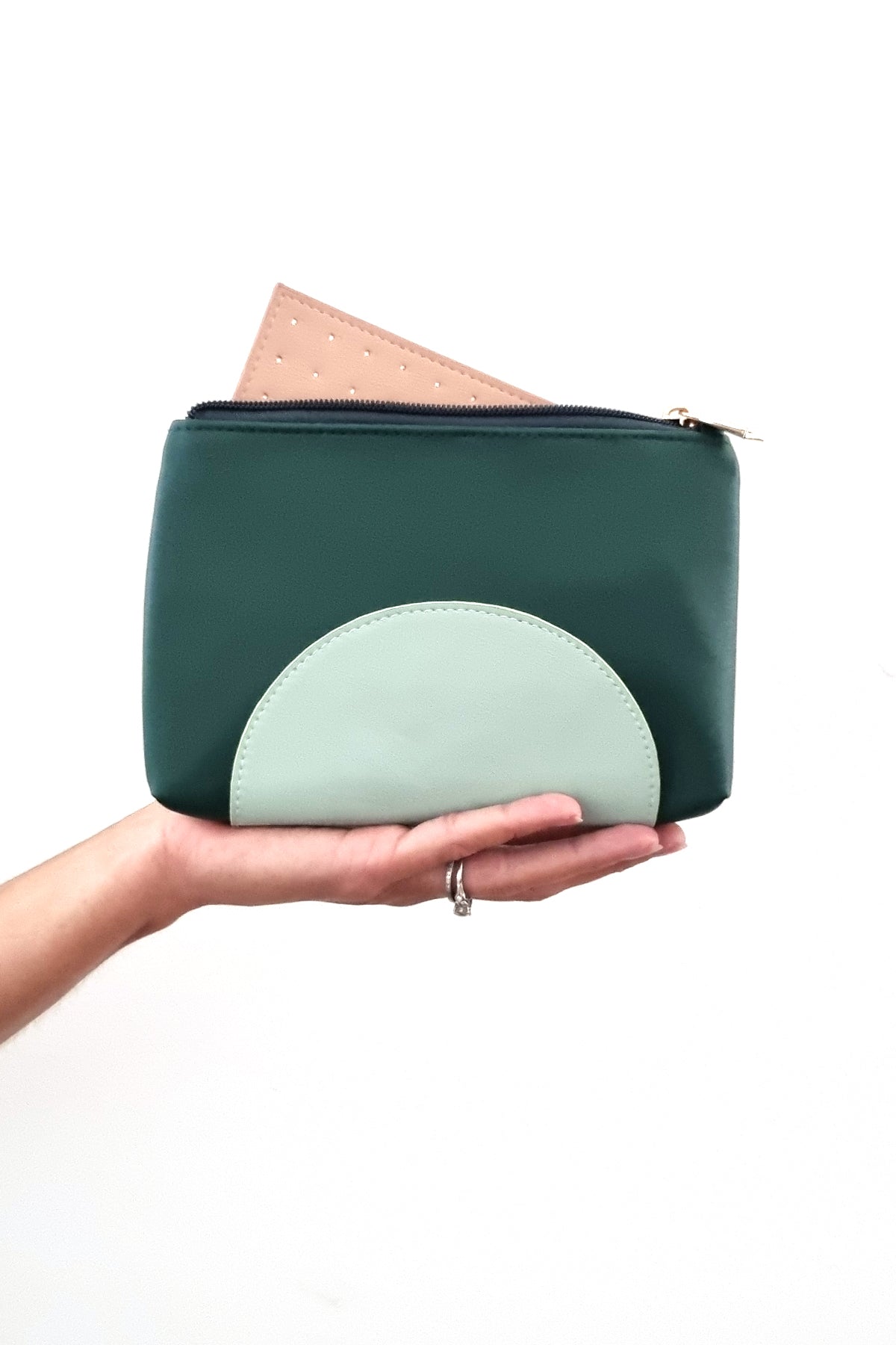 An outstretched hand holds a forest green purse against a white background. The purse has a duckegg half circle on the bottom and a gold zip, it has a peach perforated insert poking out the top of the open zip.