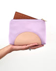 An outstretched hand holds a lilac purse against a white background. The purse has a peach half circle on the bottom and a gold zip, it has a tan perforated insert poking out the top of the open zip.