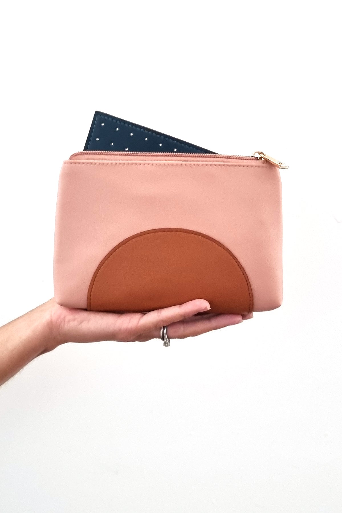 An outstretched hand holds a pink purse against a white background. The purse has a tan half circle on the bottom and a gold zip, it has a forest green perforated insert poking out the top of the open zip.