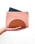 An outstretched hand holds a pink purse against a white background. The purse has a tan half circle on the bottom and a gold zip, it has a forest green perforated insert poking out the top of the open zip.