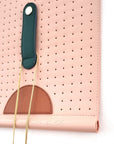 A close up image shows the detail of the wall hanging in peach. It shows the detail of the elongated forest green oval with gold button clasp from which the chain of a necklace hangs. It shows the small tan half circle, and the embossed script that says middlechild on the bottom edge of the wall hanging.