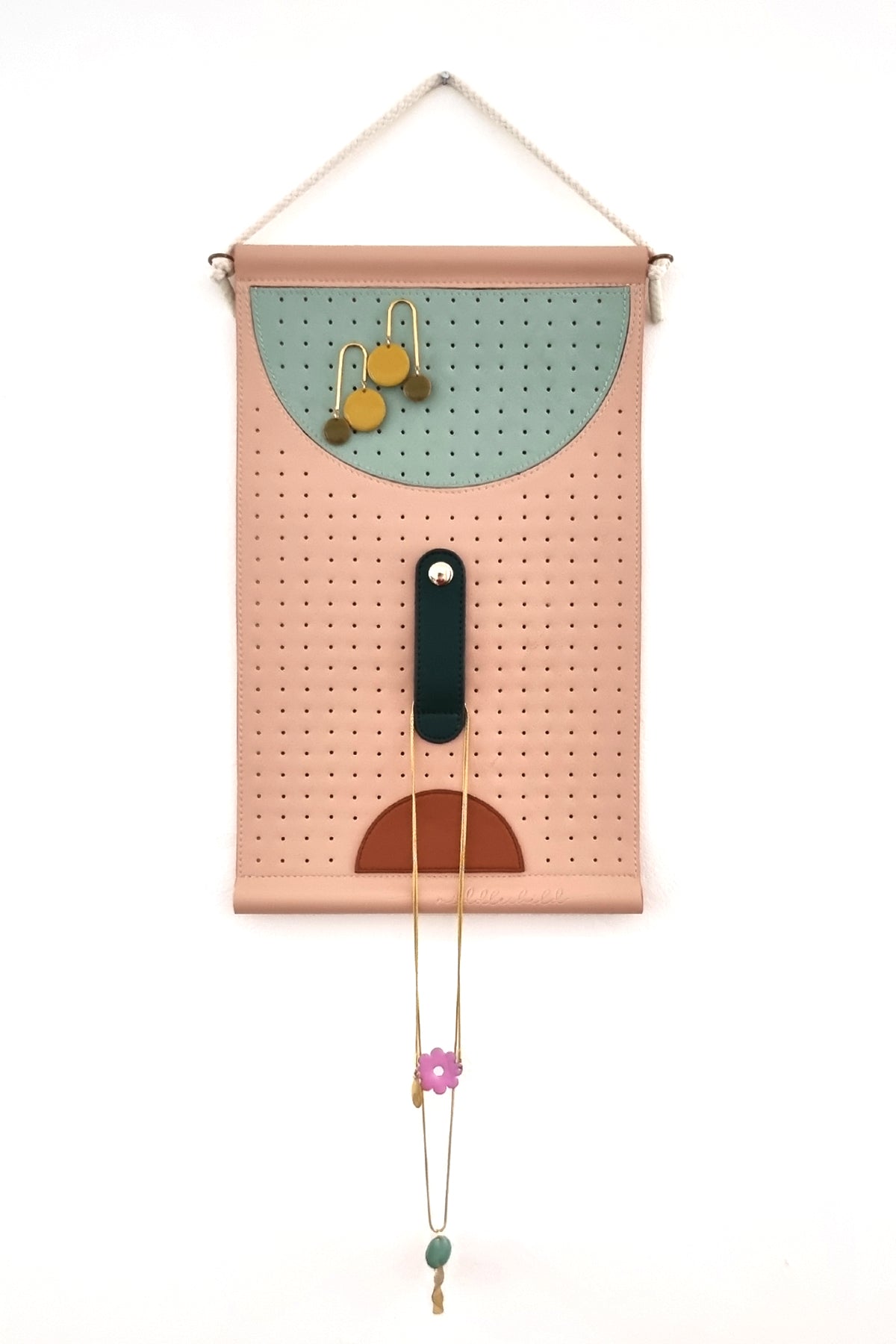 A peach perforated wall hanging is hung by a rope against a white wall. The wall hanging features a large perforated sage half circle at the top from which a pair of chartreuse earrings hang. Below this is a forest green elongated oval stitched to the middle with a gold button clasp, from this a couple of necklaces are fastened. Below this is a tan half circle.