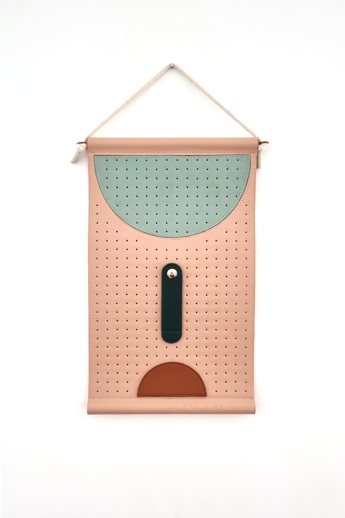 A peach perforated wall hanging is hung by a rope against a white wall. The wall hanging features a large perforated sage half circle at the top, a forest green elongated oval stitched to the middle with a gold button clasp, and below is a tan half circle.