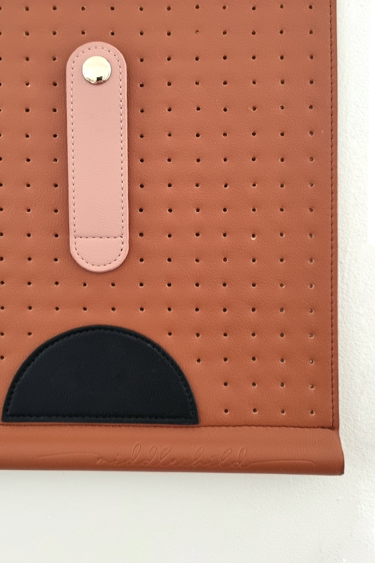 A close up image shows the detail of the wall hanging in tan. It shows the detail of the elongated peach oval with button clasp and stitch detail, the small black half circle, and the embossed script that says middlechild on the bottom edge of the wall hanging.