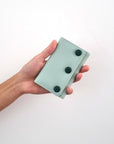 An outstretched hand holds a duckegg jewellery snug against a white wall. The snug features duckegg PU and has three PU covered buttons in teal.