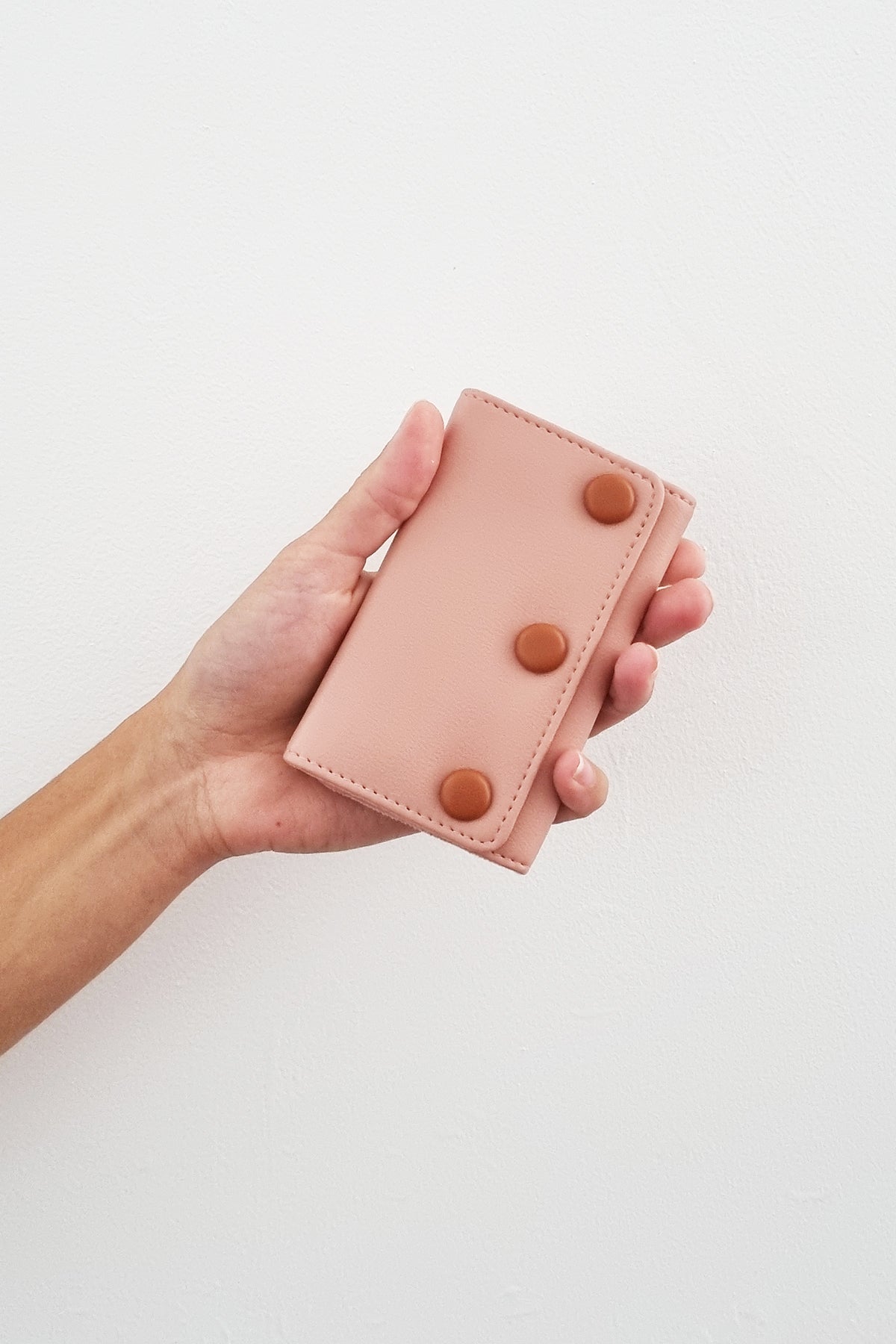 An outstretched hand holds a pink jewellery snug against a white wall. The snug features pink PU and has three PU covered buttons in tan.