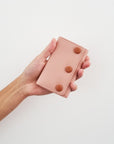 An outstretched hand holds a pink jewellery snug against a white wall. The snug features pink PU and has three PU covered buttons in tan.