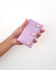 An outstretched hand holds a lilac jewellery snug against a white wall. The snug features purple PU and has three PU covered buttons in pink.