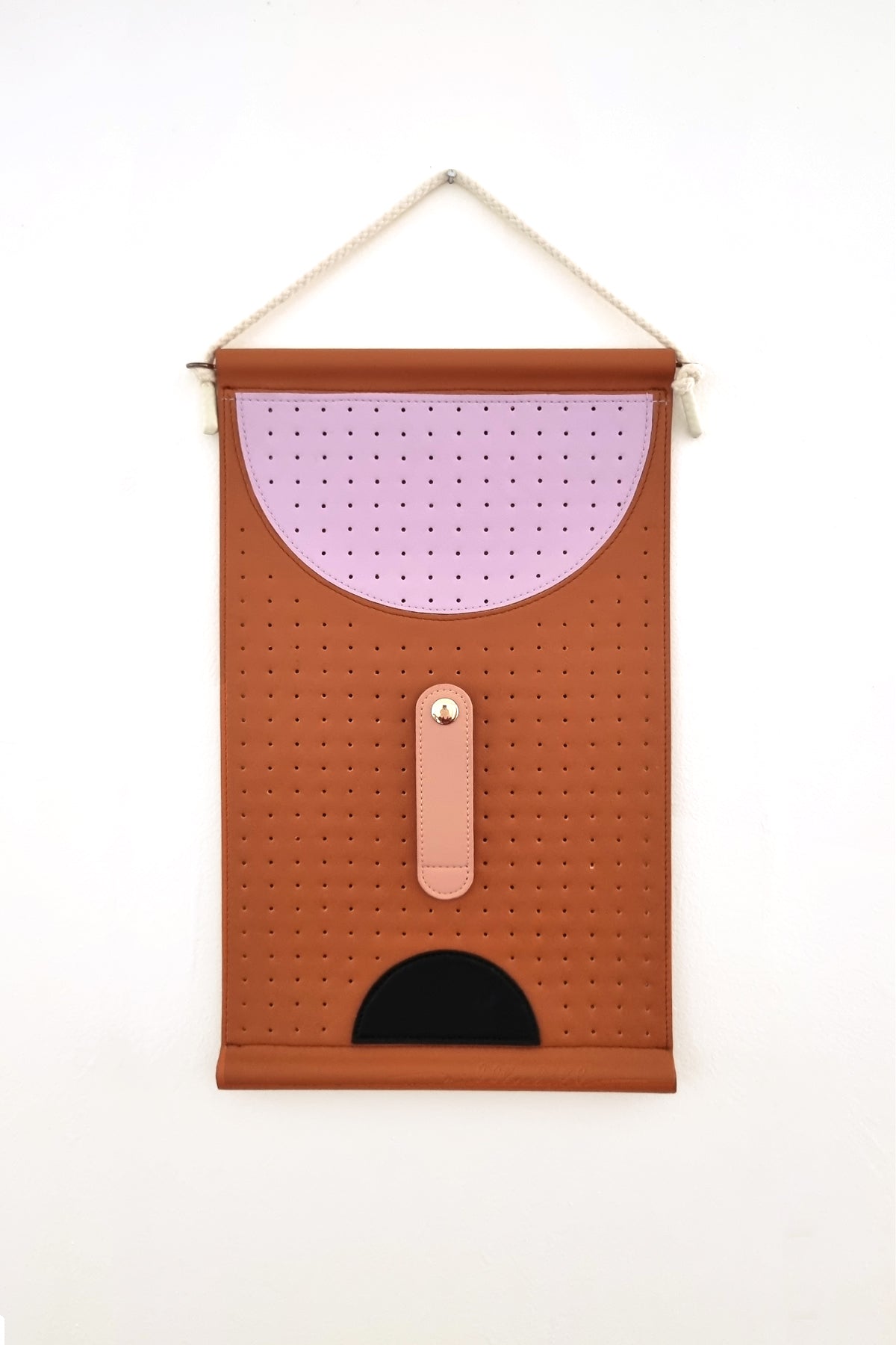 A perforated tan wall hanging is hung by a rope against a white wall. The wall hanging features a large perforated lilac half circle at the top, a peach elongated oval stitched to the middle with a gold button clasp, and below is a black half circle. 