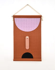 A perforated tan wall hanging is hung by a rope against a white wall. The wall hanging features a large perforated lilac half circle at the top, a peach elongated oval stitched to the middle with a gold button clasp, and below is a black half circle. 
