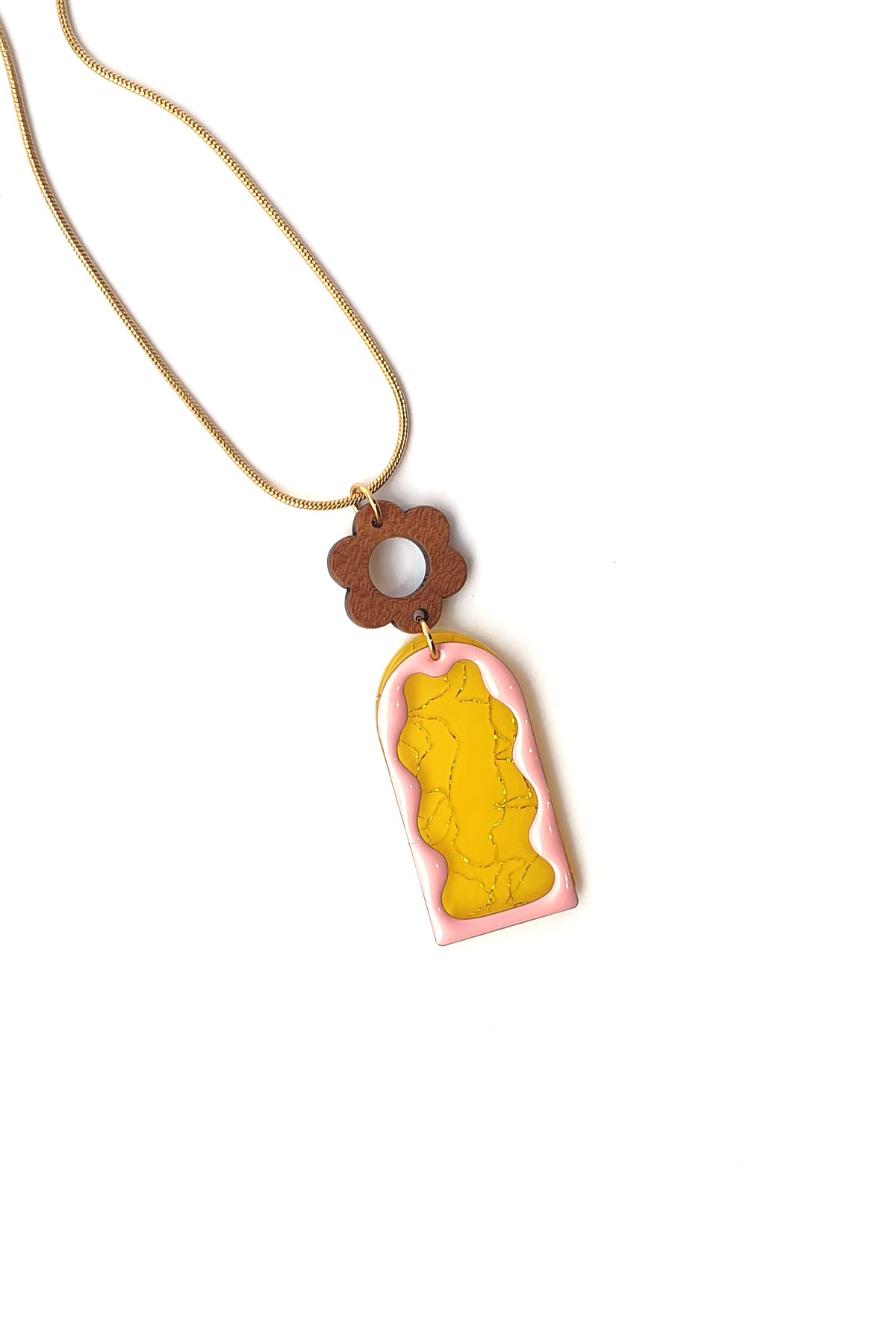 A necklace lays against a white background. It features a gold chain, a wooden flower top, followed by a yellow acrylic arch with silver thread detail overlaid by a pink acrylic wavy arch frame.