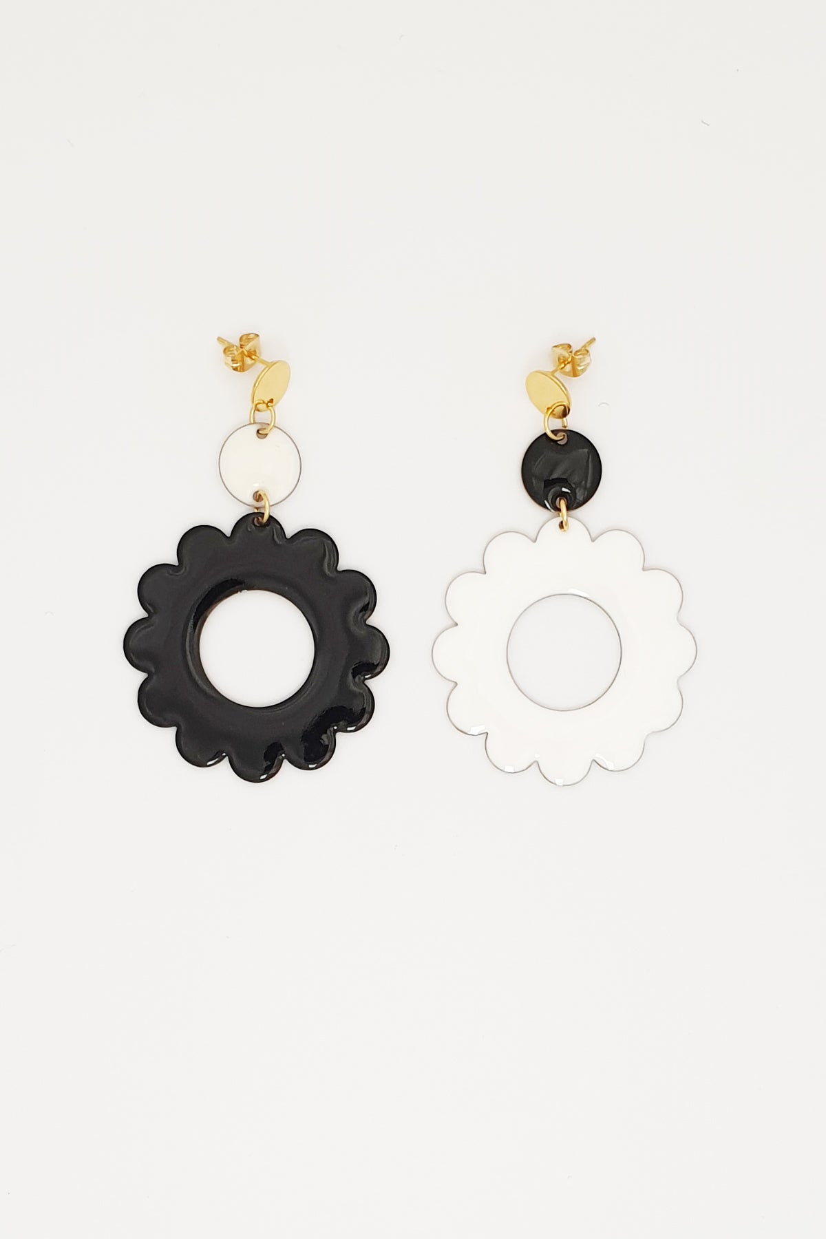 A pair of stud dangle earrings lay on white background. The left earring features a white enamel dot connected to a larger black enamel flower. The right earring is flipped and features an enamel black dot connected to a larger white enamel flower 