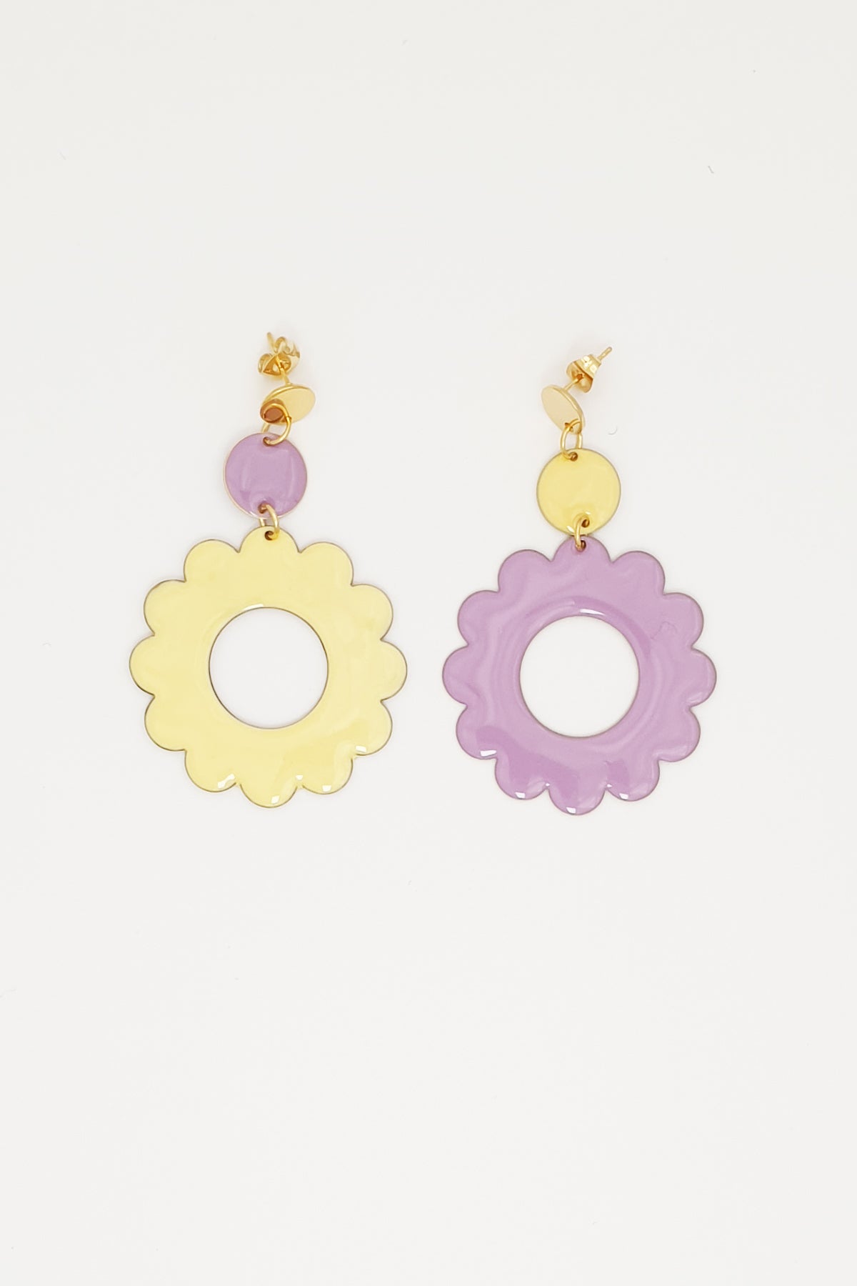 A pair of stud dangle earrings lay on white background. The left earring features a lilac enamel dot connected to a larger enamel lemon flower. The right earring is flipped and features an enamel lemon dot connected to a larger lilac enamel flower 
