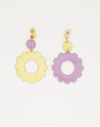 A pair of stud dangle earrings lay on white background. The left earring features a lilac enamel dot connected to a larger enamel lemon flower. The right earring is flipped and features an enamel lemon dot connected to a larger lilac enamel flower 