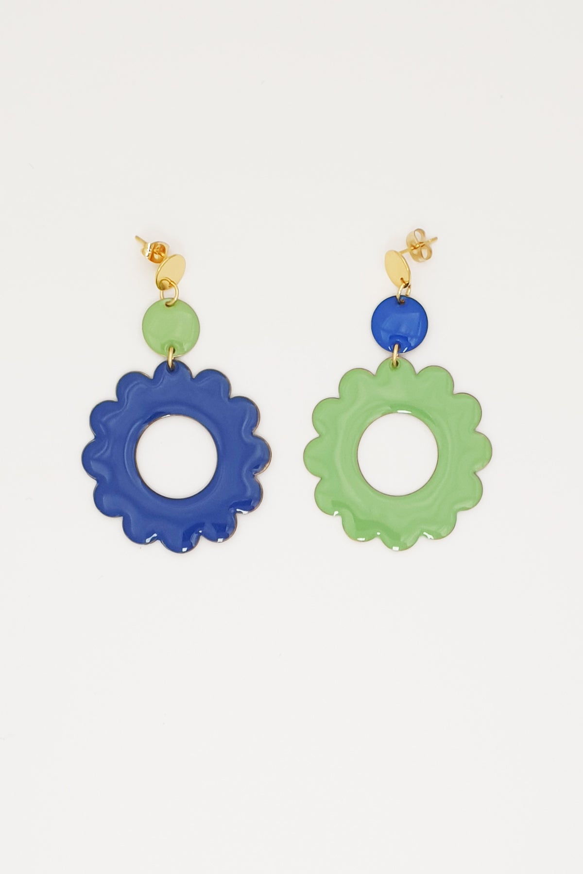 A pair of stud dangle earrings lay on white background. The left earring features a sage enamel dot connected to a larger sapphire blue enamel flower. The right earring is flipped and features an enamel sapphire blue dot connected to a larger sage enamel flower.
