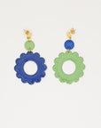 A pair of stud dangle earrings lay on white background. The left earring features a sage enamel dot connected to a larger sapphire blue enamel flower. The right earring is flipped and features an enamel sapphire blue dot connected to a larger sage enamel flower.