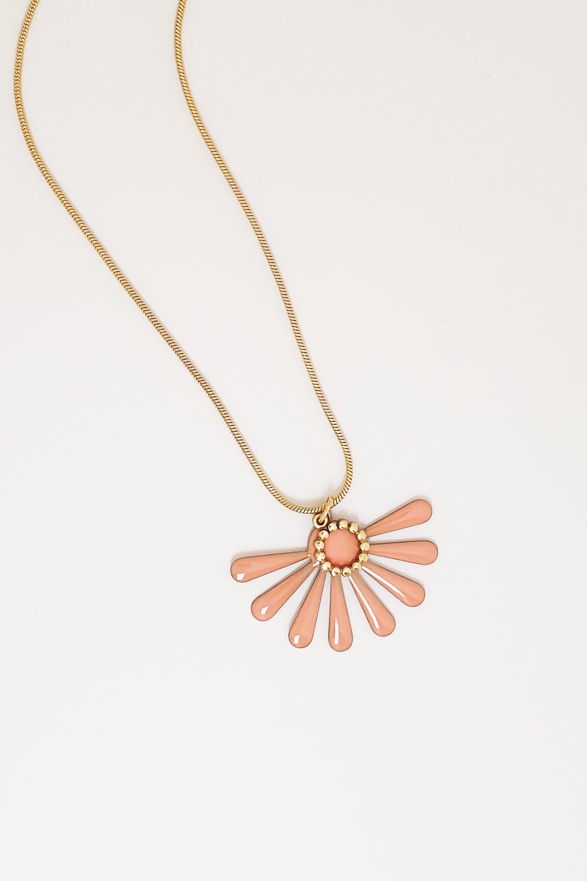 A necklace sits against a white background. It features a gold chain and an apricot coloured floral enamel shape. A tiny brass ring encircles the disk of the floral enamel piece.