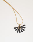 A necklace sits against a white background. It features a gold chain and a black coloured floral enamel shape. A tiny brass ring encircles the disk of the floral enamel piece.