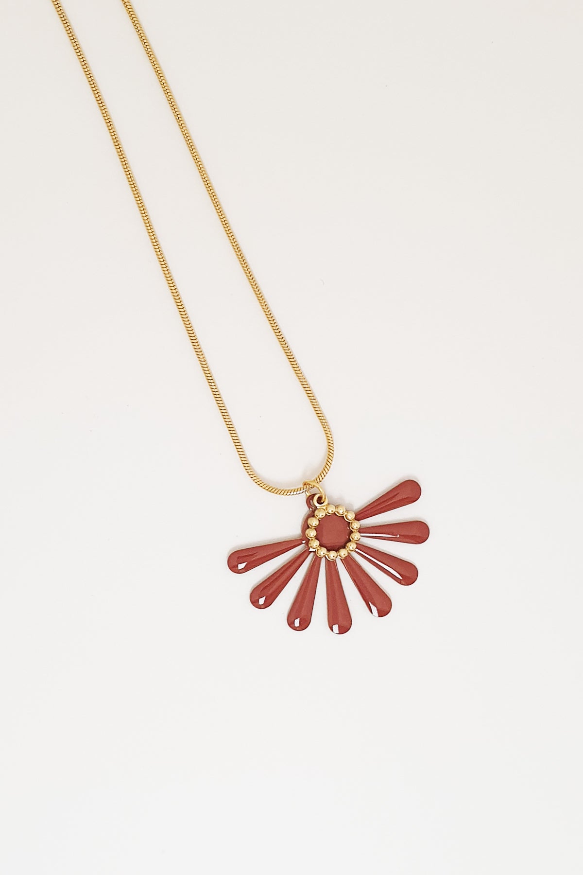 A necklace sits against a white background. It features a gold chain and a brown coloured floral enamel shape. A tiny brass ring encircles the disk of the floral enamel piece.