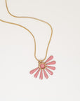 A necklace sits against a white background. It features a gold chain and an pink coloured floral enamel shape. A tiny brass ring encircles the disk of the floral enamel piece.