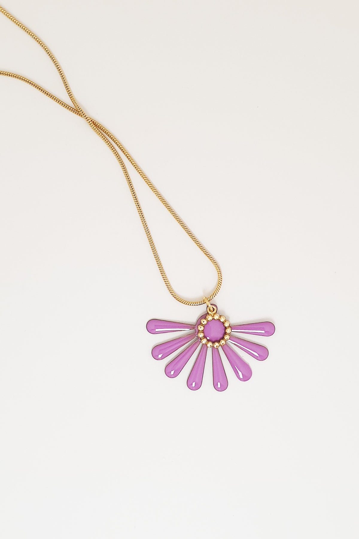 A necklace sits against a white background. It features a gold chain and an orchid pink coloured floral enamel shape. A tiny brass ring encircles the disk of the floral enamel piece.