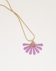 A necklace sits against a white background. It features a gold chain and an orchid pink coloured floral enamel shape. A tiny brass ring encircles the disk of the floral enamel piece.
