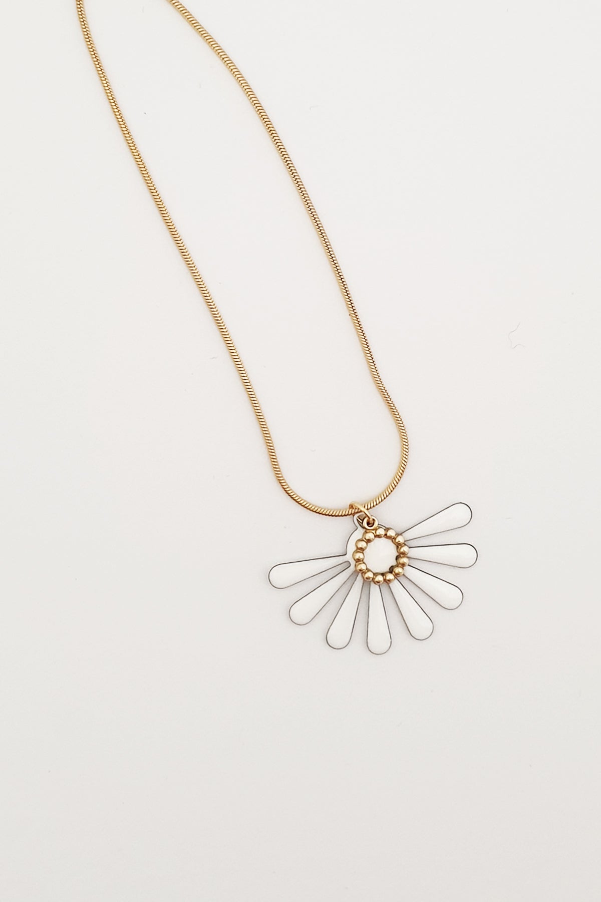 A necklace sits against a white background. It features a gold chain and a white coloured floral enamel shape. A tiny brass ring encircles the disk of the floral enamel piece.