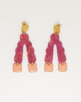 A pair of stud dangle earrings sit against a white background. They feature a textured circular gold stud top, a magenta wavy upside down V-shaped acrylic, and peach square shaped enamel drops hanging from each end of the acrylic.