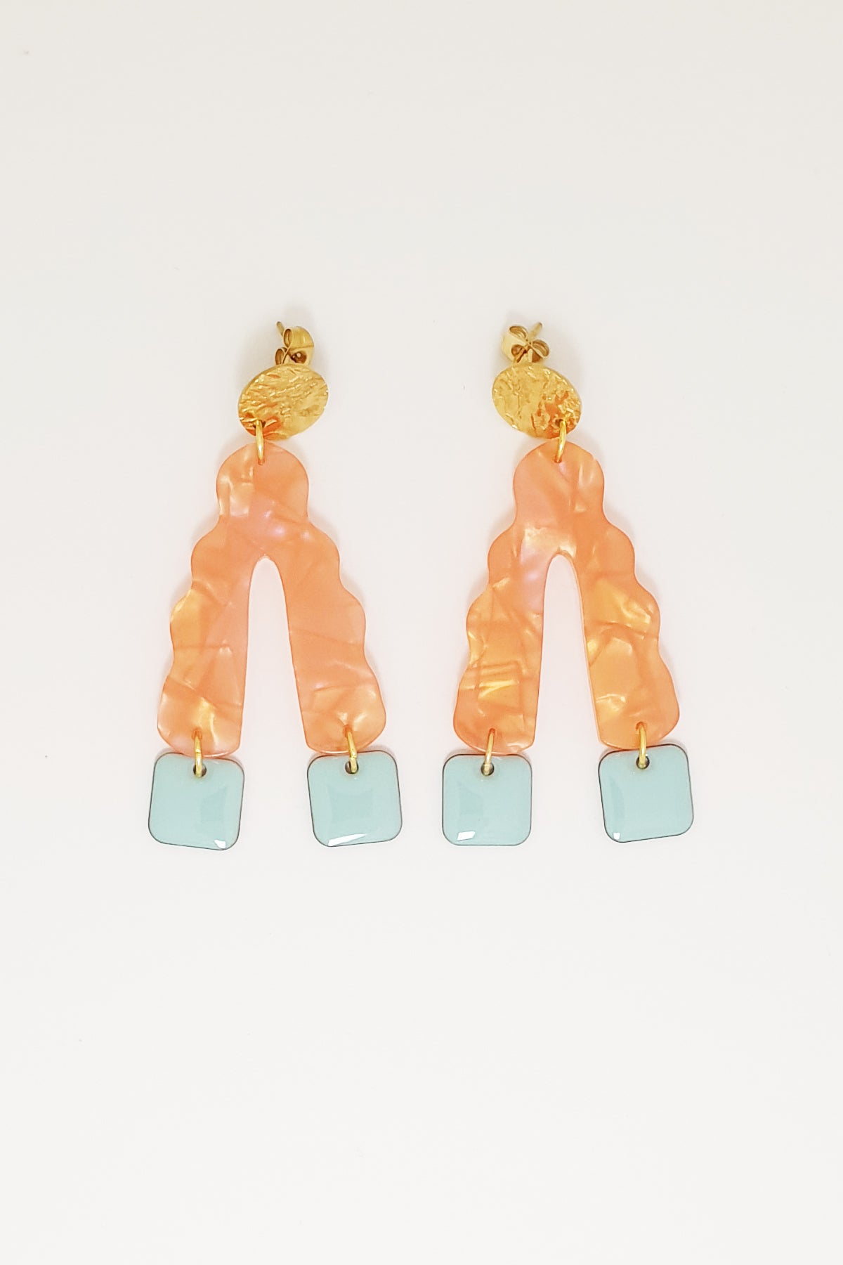 A pair of stud dangle earrings sit against a white background. They feature a textured circular gold stud top, an orange wavy upside down V-shaped acrylic, and honeydew blue square shaped enamel drops hanging from each end of the acrylic.