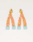 A pair of stud dangle earrings sit against a white background. They feature a textured circular gold stud top, an orange wavy upside down V-shaped acrylic, and honeydew blue square shaped enamel drops hanging from each end of the acrylic.