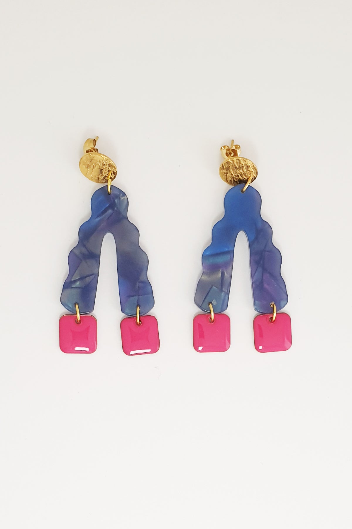 A pair of stud dangle earrings sit against a white background. They feature a textured circular gold stud top, a blue wavy upside down V-shaped acrylic, and magenta square shaped enamel drops hanging from each end of the acrylic.