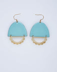 A pair of statement earrings with a hook sit against a white background. They feature a sky blue enamel arch and a horizontal plated brass D shape with scalloped detail.