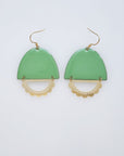 A pair of statement earrings with a hook sit against a white background. They feature a sage green enamel arch and a horizontal plated brass D shape with scalloped detail.